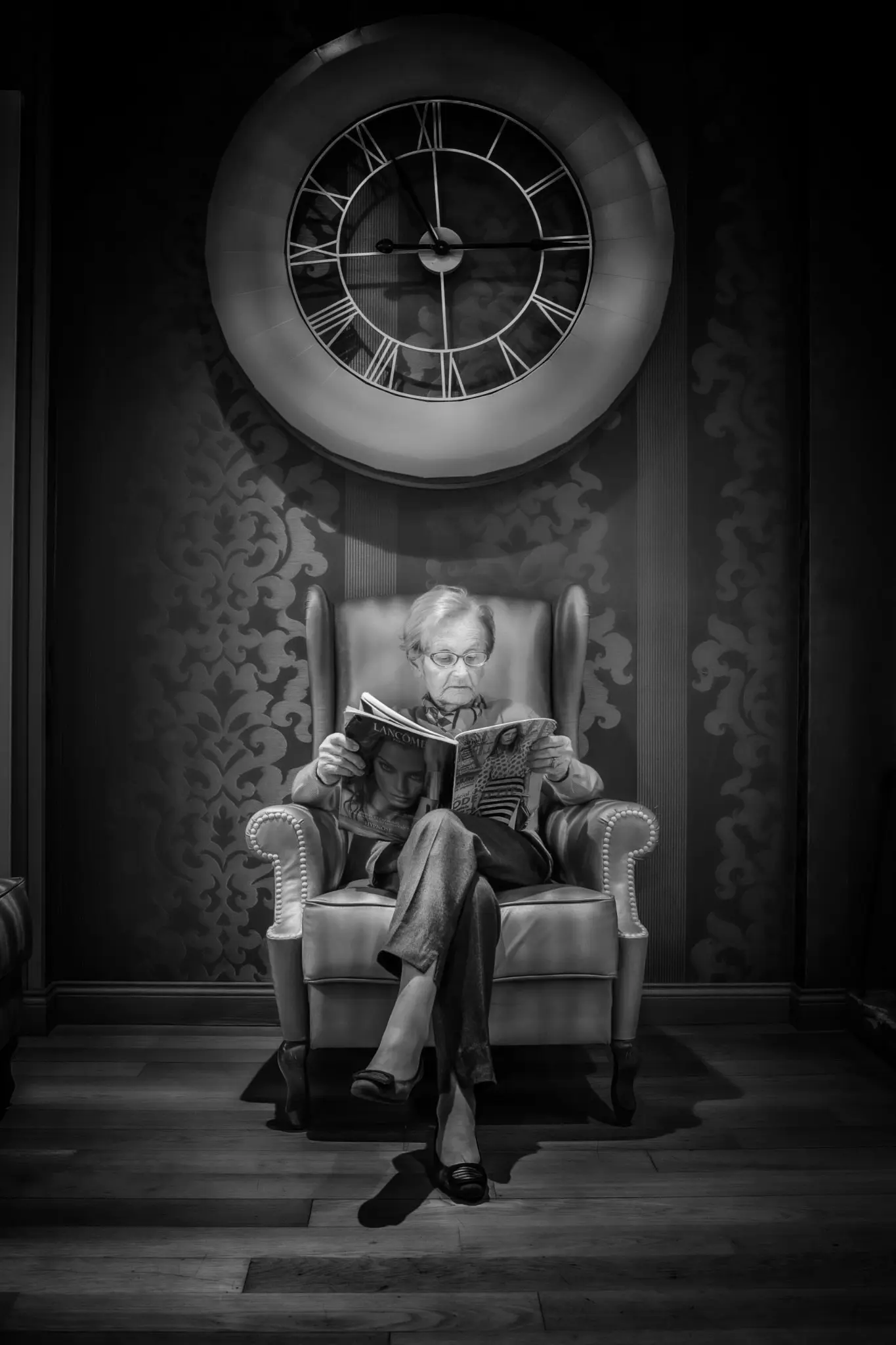 Une femme âgée assise dans un fauteuil sous une grande horloge, photographiée par Didier Carluccio