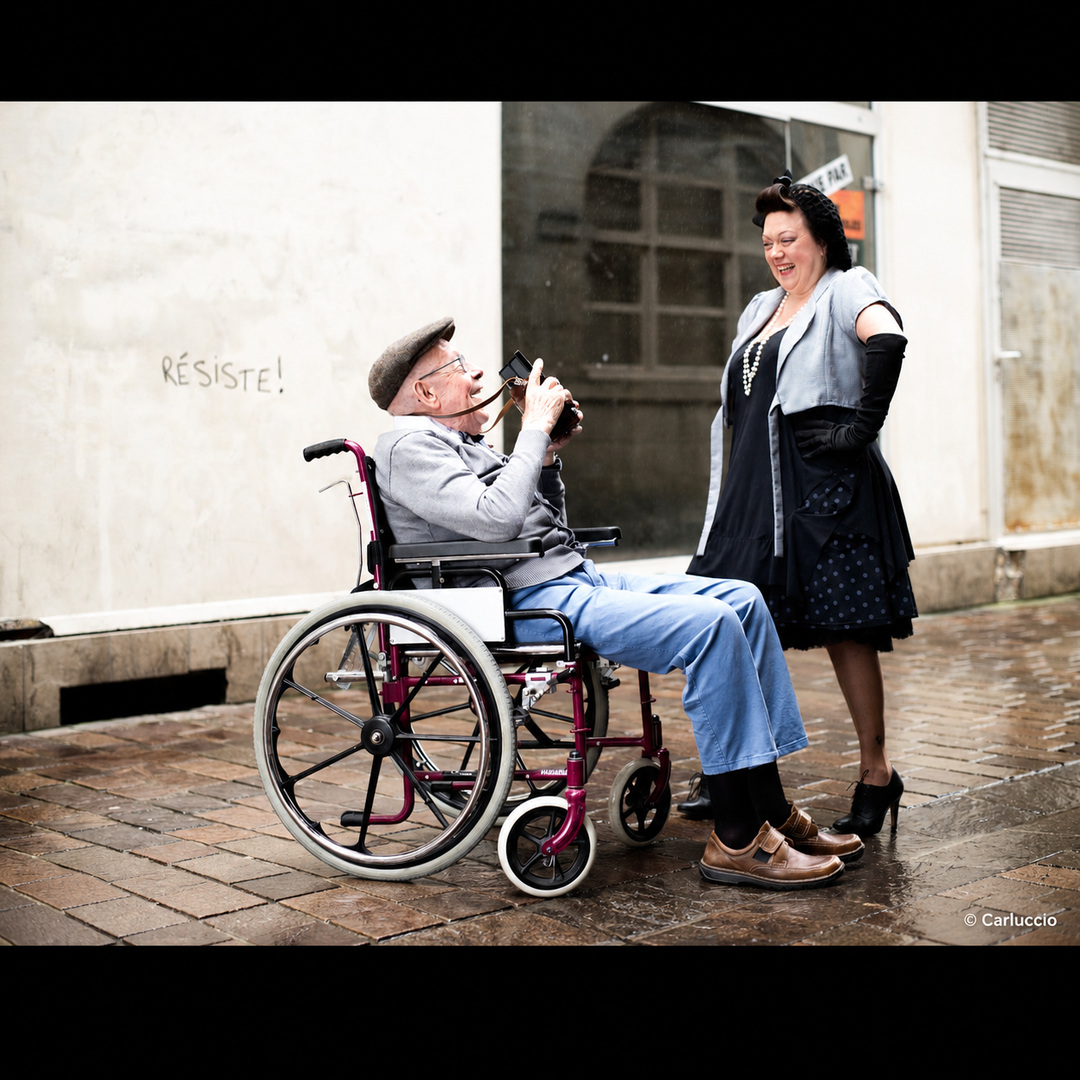 Un homme âgé en fauteuil roulant tient un vieil appareil photo devant son visage et photographie une femme qui pose face à lui en riant, lors d'une séance en extérieur.