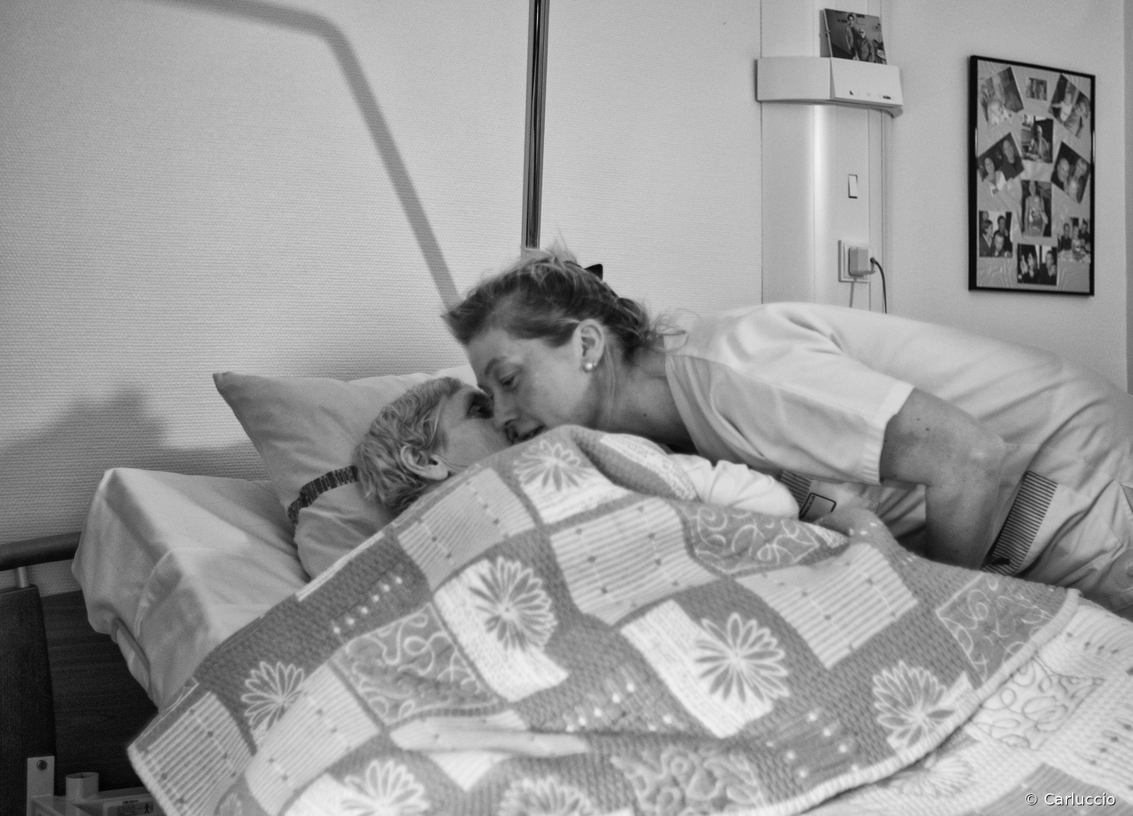 Le bisou du soir Photographie en noir et blanc d’une soignante penchée pour embrasser une résidente allongée sous une couverture à motifs. Des photographies personnelles sont accrochées au mur de la chambre.