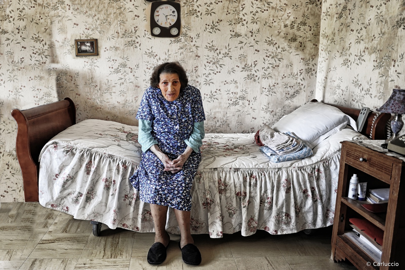 Un peu de chez soi Une résidente assise au bord d’un lit ancien en bois, les mains jointes sur les genoux. Sa blouse, le couvre-lit et la tapisserie sont ornés de motifs fleuris.