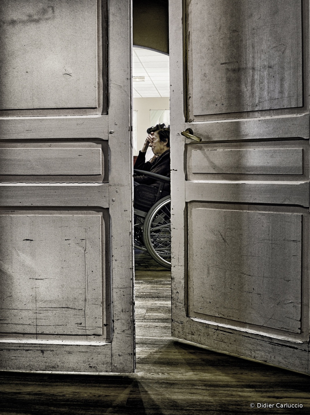Entre les portes À travers deux grandes portes ouvertes, une personne âgée assise seule dans un fauteuil apparaît au fond d’un salon, petite silhouette dans un vaste espace.