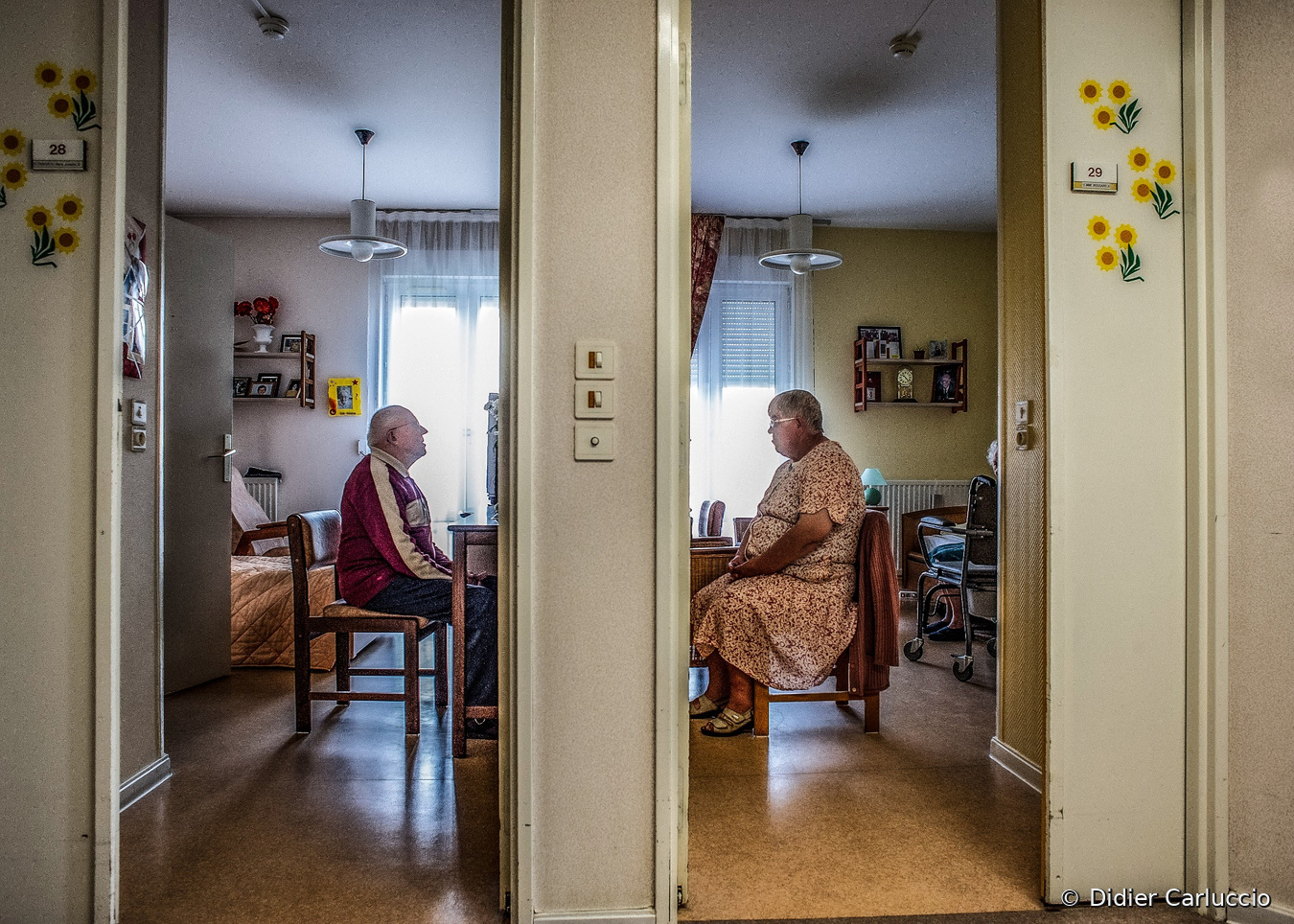 Photographie documentaire de Didier Carluccio montrant deux résidents âgés assis dans leurs chambres respectives, portes ouvertes, presque face à face. Une cloison centrale sépare leurs espaces de vie, décorés de photographies et d'objets personnels.