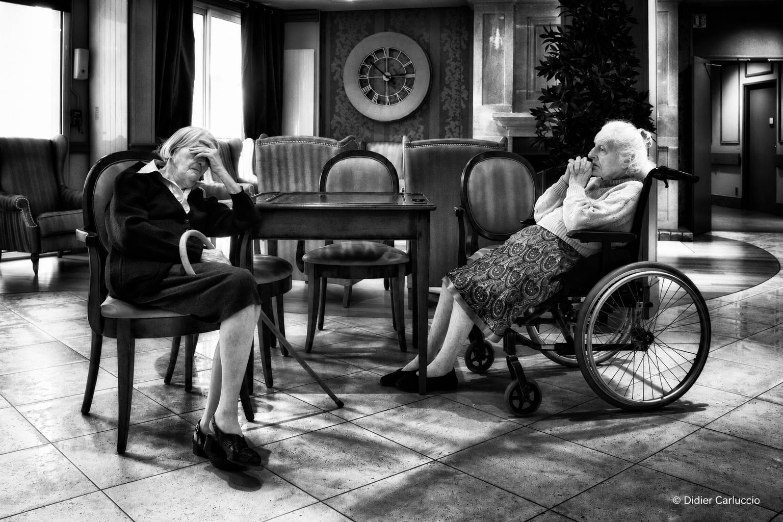 Photographie en noir et blanc de deux femmes âgées assises dans un espace d’accueil. L’une est sur une chaise, la main sur le front, l’autre dans un fauteuil roulant, les mains près du visage. Une grande horloge est visible derrière elles.