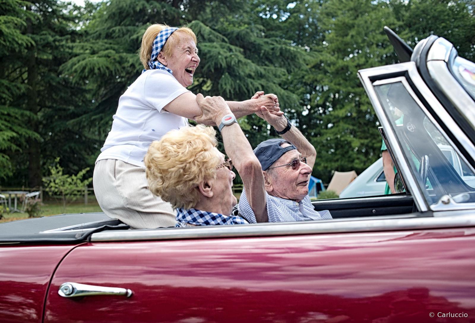 Une femme âgée rit debout à l'arrière d'un cabriolet ancien rouge, entourée de deux autres résidents, lors d'une fête organisée en extérieur.